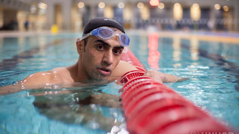 Hussein está em uma piscina segurando a raia vermelha, a cabeça está fora da água e ele olha para a câmera, os óculos estão na testa.