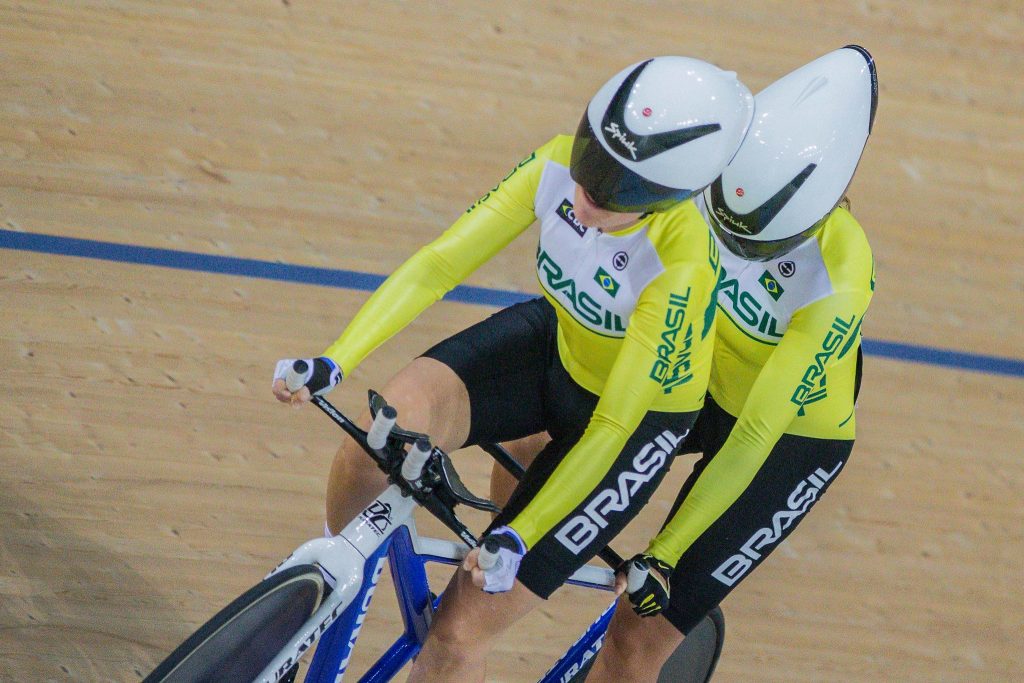 2 atletas competindo no ciclismo em cima de uma bicicleta com 2 bancos, usando capacetes brancos, representando o Brasil com uma camisa amarela e branca.