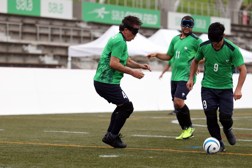 Três jogadores homens presentes em um gramado sintético, usam venda preta de futebol de 5 nos olhos, na disputa por uma só coisa: a bola