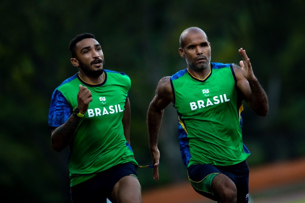 Foto de dois homens negros, ao lado esquerdo está o guia Wendel Souza, ele tem cabelo curto preto e barba. Ao lado dele, ligados por uma corda na mão esquerda de Wendel e direita de Daniel, o velocista, careca, correm. ambos vestem um uniforme verde da seleção brasileira e aparentam estar concentrados. 