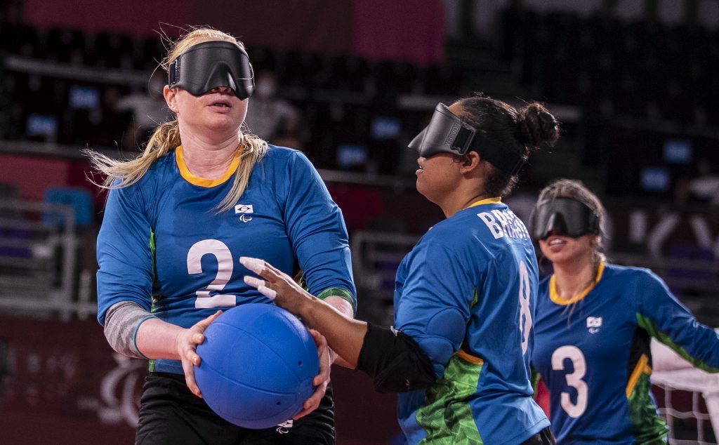 Foto das jogadoras Ana Gabrielly e Jéssica Vitorino, uma mulher branca e loira e outra mulher negra, durante partida de golbol. Elas vestem o uniforme azul da seleção brasileira. A jogadora branca está com uma bola azul nas mãos e a outra jogadora segura a bola com sua mão esquerda. 