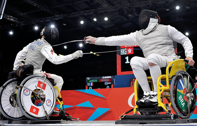 Foto de arena com dois lutadores de esgrima sobre cadeira de rodas, um apontando uma longa espada pontiaguda sobre o outro. O primeiro tem cadeira de rodas com símbolo da Inglaterra, o segundo, do Brasil.