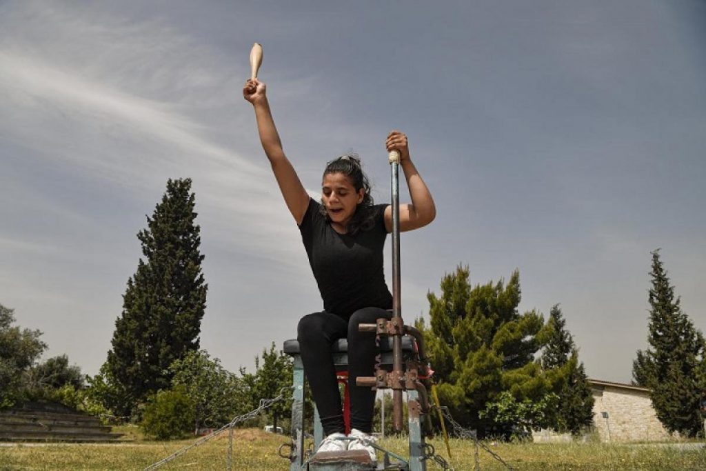 Alia está em um lugar aberto, sentada em um banco de ferro, levantando um arco com seu braço direito e uma barra de ferro com o esquerdo.