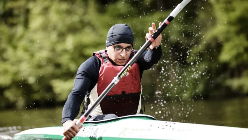 Annas, um homem branco, está em uma canoa praticando remo com o todo coberto com o uniforme preto de nado.