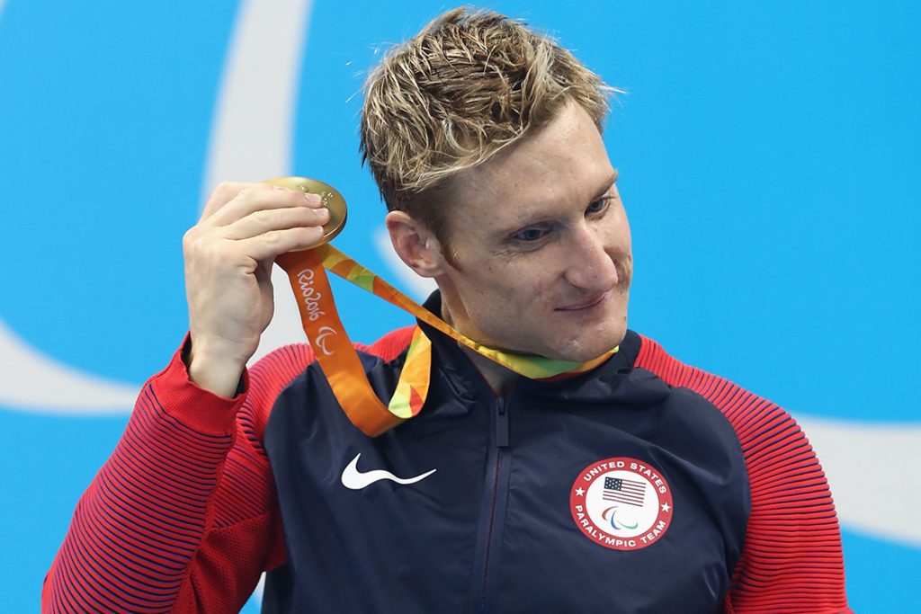 Atleta branco de cabelo loiro americano com blusa representando seu país. Ele está dentro do pódio e ergue sua medalha na altura do ouvido para escutá-la