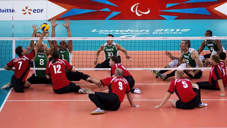 Foto mostra dois times de volei em quadra, todos sentados, disputando uma bola durante partida