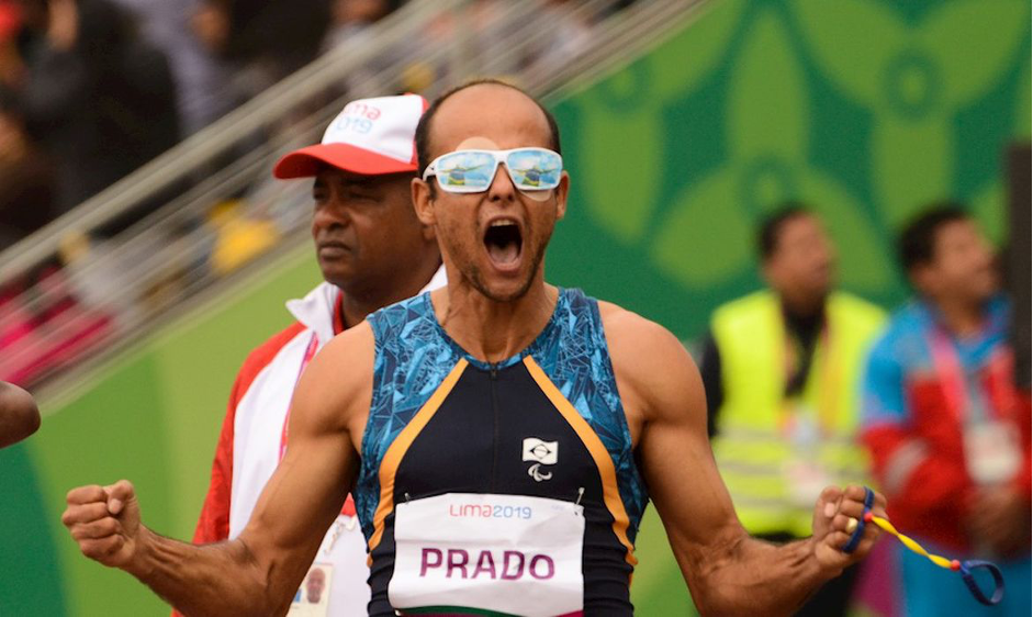 Atleta Lucas Prado, homem negro, calvo, usando óculos e uma camisa azul sem mangas comemora ao final de uma prova. O atleta está num estádio e ao fundo outras pessoas acompanham o momento.