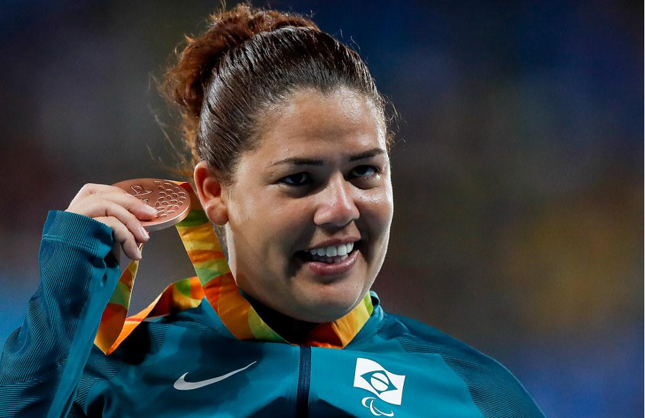 Atleta Marivana Oliveira da Nóbrega, mulher branca, cabelos castanhos, vestida com um casaco azul marinho, sorri enquanto segura com a mão direita a medalha de bronze. O fundo da imagem está desfocado. Foto: CPB