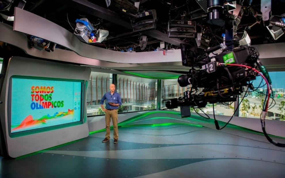 Fotografia de um estudio de televisão. À esquerda há um telão onde aperece a mensagem "somos todos olímpicos". Ao lado do telão está um apresentador de TV. Na lateral direita é há uma câmera de vídeo filmando o jornalista.