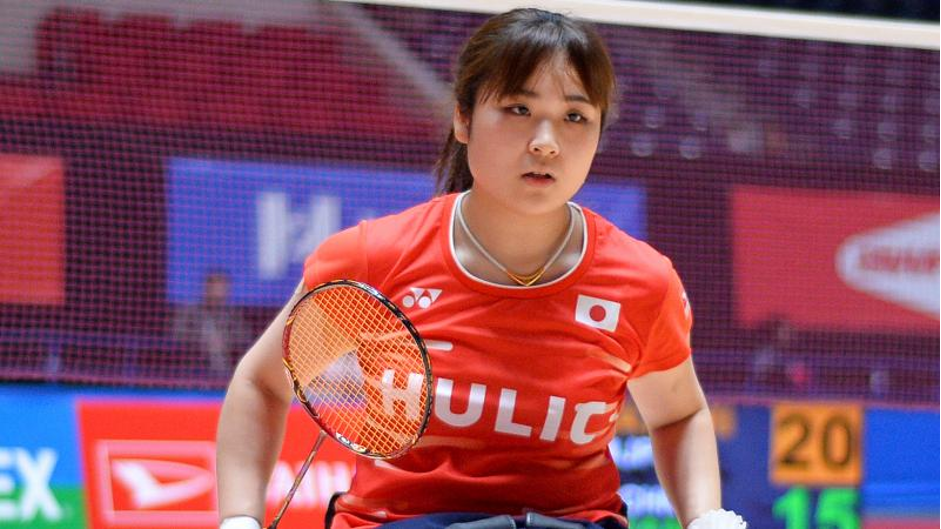 Foto de Sarina em uma partida de ParaBadminton. Tem cabelo castanho longo, usa uma franja, está com um colar branco, segura uma raquete e está vestida com uma camisa vermelha com a bandeira do Japão.