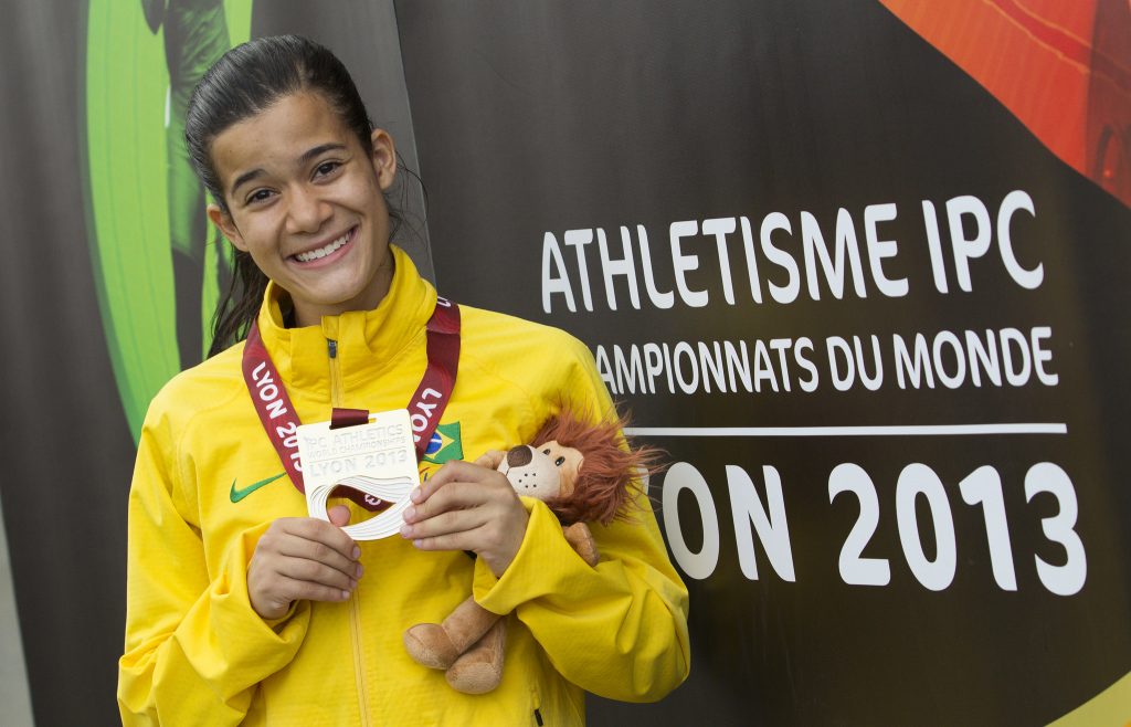 Verônica está com o cabelo preso em um rabo de cavalo e usando o uniforme, uma jaqueta amarela com a bandeira do Brasil. Ela sorri para a câmera e com as duas mãos segura a medalha de ouro que está pendurada em volta de seu pescoço. Com o braço esquerdo, ela segura junto ao corpo, o mascote de pelúcia do campeonato, um leão.