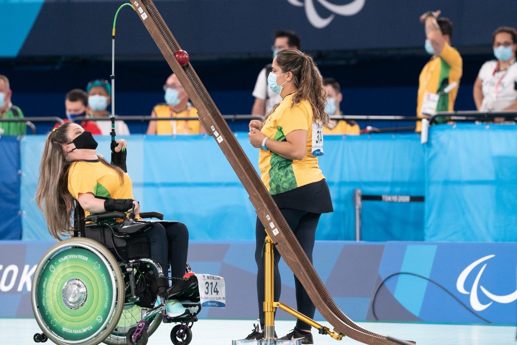 Foto em uma quadra onde Evani Calado, mulher branca, usa um uniforme amarelo com verde e leggins e máscara de proteção preta. Elva tem cabelo loiro preso em uma xuxinha verde e amarela. Está em uma cadeira de rodas e na sua frente uma calha de madeira está com uma bolinha vermellha. ela olha pra cima e ergue uma vara de alumnio presa em sua mão pra cima onde a bolinha vermelha está. ao lado está a calheira Renata, de costas para o jogo, com uo uniforme da seleção brasileira também.
