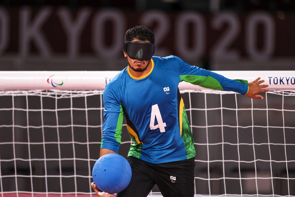 Foto de Leomon, o camisa 4, no centro do gol do golbol durante uma partida. Ele carrega a bola azul e se prepara para lançar. Ele veste a venda nos olhos e o uniforme azul da seleção brasileira