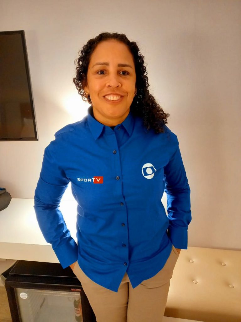 Adria posa para a foto de perfil. Ela veste camisa azul da equipe de comentaristas da emissora e calça beje. Tem os cabelos soltos cacheados e sorri.