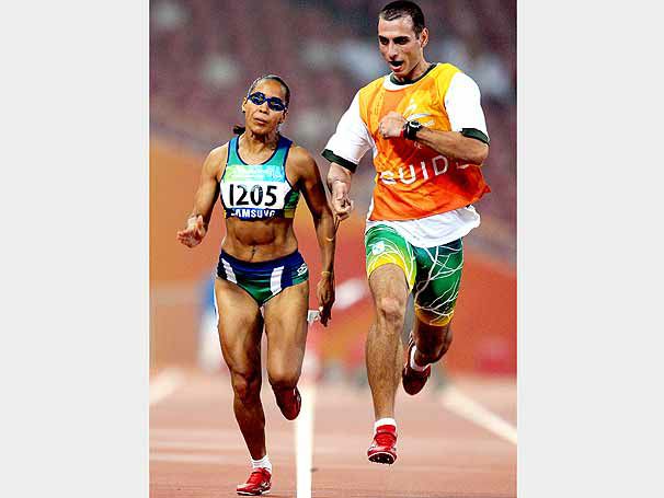 na imagem Adria - a esquerda - corre de frente , em uma pista de atletismo, acompanhada pelo atleta guia - a direita. Ela tem a pele parda, cabelos escuros e presos. Veste um top e shorts curto.