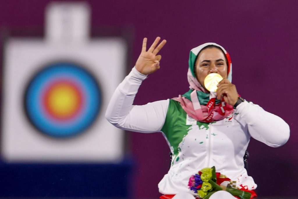 Zahra cobre seus cabelos com uma burca, beija sua medalha. Atrás dela há um painel com alvo do tiro com arco.