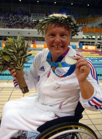 Beatrice está sobre cadeira de rodas, sorrindo para a camera e mostrado sua medalha. Ela está em um ginásio com piscinas e segura ramo de folhas de louro na mão e em coroa na cabeça.