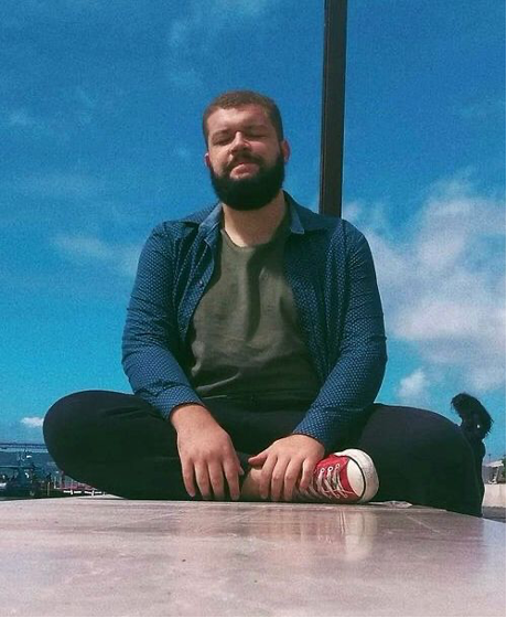 Emerson, homem branco, cabelo castanho raspado, barba curta preta. Usa uma camiseta verde e por cima uma camisa manga longa azul com bolinhas, calça de moletom preta e all star vermelho com branco. Está sentado em uma mureta, aparece de frente, com a perna direita cruzada na frente, segurando com as mãos, ao fundo, céu azul limpo.