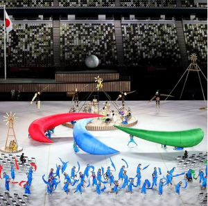 foto da cerimônia de abertura da Paralimpíada onde está mostrando uma performance artística feita por várias pessoas. No centro do palco, os artistas carregam três arcos de espuma em vermelho, verde e azul que juntos formam o “Agitos”, símbolo paralimpíco.