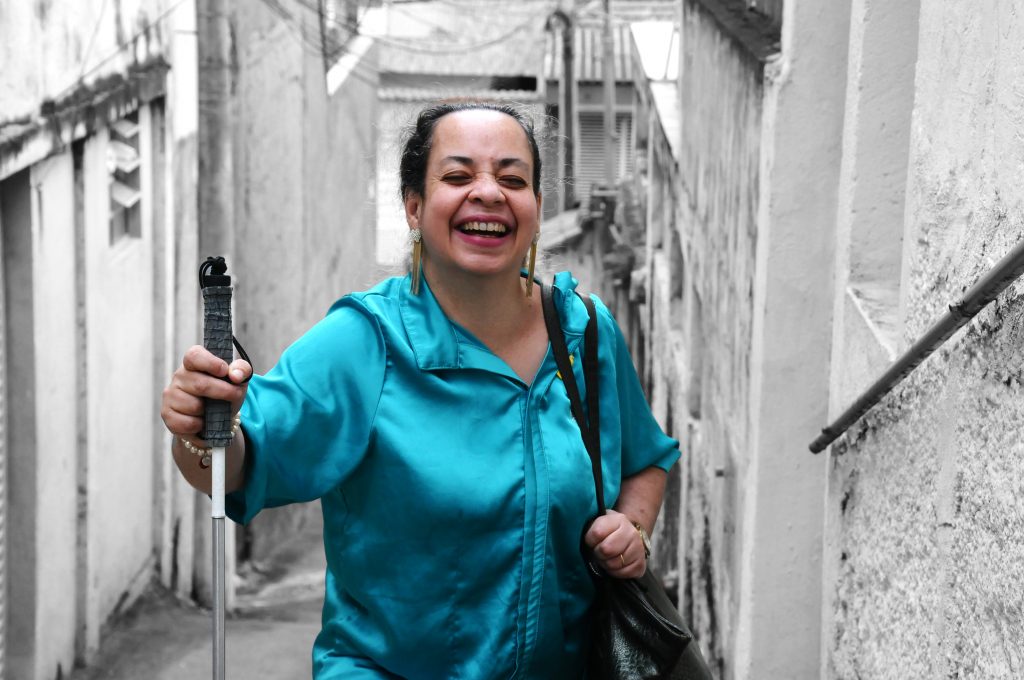 Foto colorida da cintura para cima de Regina Célia, mulher branca, de 53 anos, sorrindo. Segura bengala com a mão direita, no alto de uma ladeira. Ao fundo, casas em preto e branco. Veste blusa azul esverdeada cintilante. Usa cabelo preso, batom, brincos, bolsa preta, pulseira de pérolas, relógio e aliança.