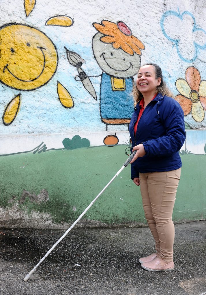Foto colorida de Regina Célia, de pé, segurando bengala. Ela sorri e veste jaqueta azul, camisa polo vermelha, calça bege e tênis rosa. Ao fundo, no muro da escola, grafite infantil de menin(e) com pincel, sol, nuvens e flor.