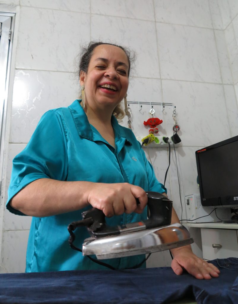 Foto colorida da cintura para cima de Regina Célia sorrindo. Na mão direita, segura ferro de passar roupa e apoia a outra sobre a calça jeans na tábua de passar. À direita, computador sobre a bancada.