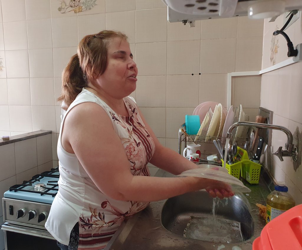 Foto colorida da cintura para cima de Ana Cristina, 51 anos, mulher branca, de perfil, lavando louça. Ela tem cabelo liso, louro escuro e preso. Veste blusa branca sem manga com estampa de flores.