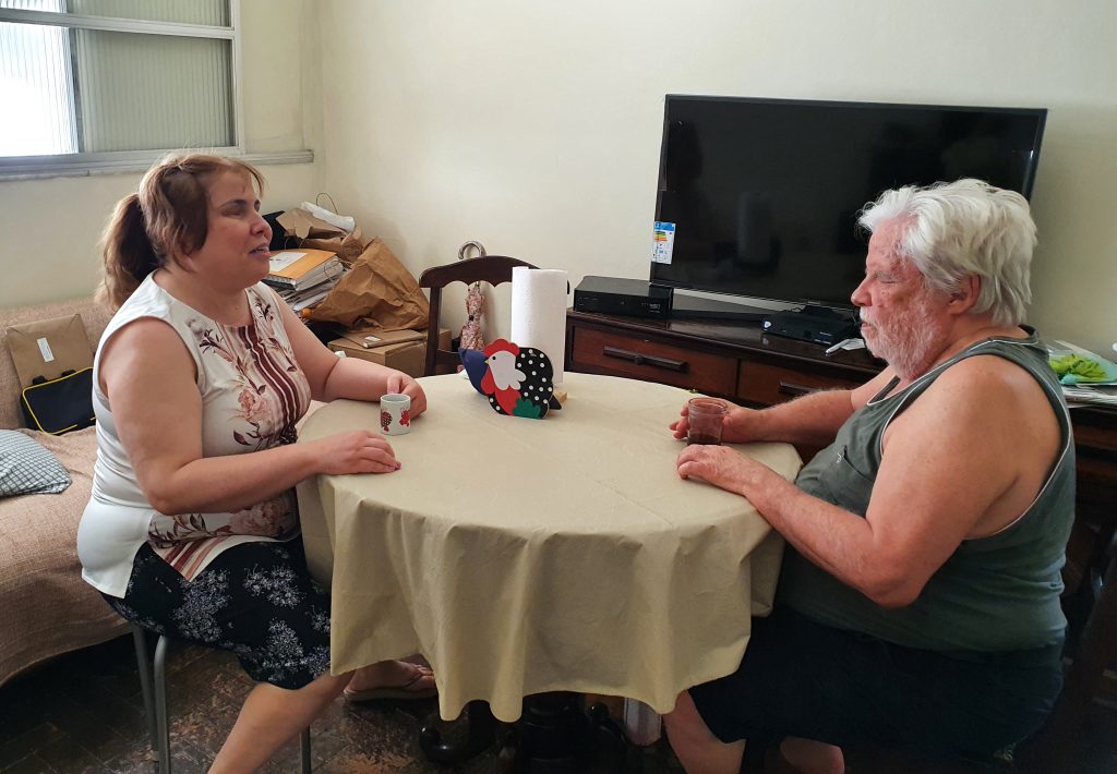 Foto colorida de Ana Cristina, mulher branca, de cabelo preso, louro escuro, liso, com o pai, de 82 anos, homem branco, de barba e cabelos brancos, sentados à mesa redonda. Ela segura uma xícara e ele um copo com café. Ao fundo, uma televisão. Ambos estão de bermuda e camiseta sem manga.
