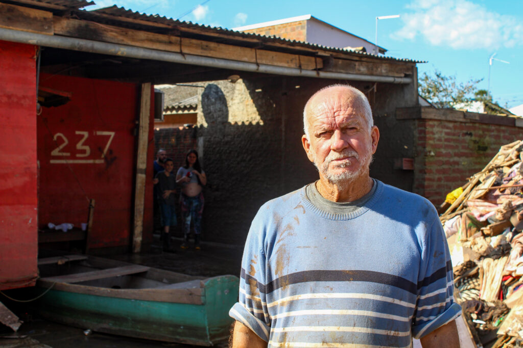 Darcy, vítima das enchentes no Rio Grande do Sul com sua casa afetada ao fundo