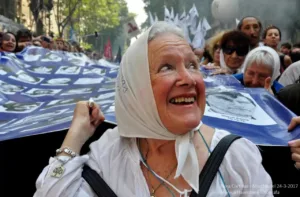 Nora Cortiñas sorri com um lenço branco na cabeça em frente a uma grande bandeira.