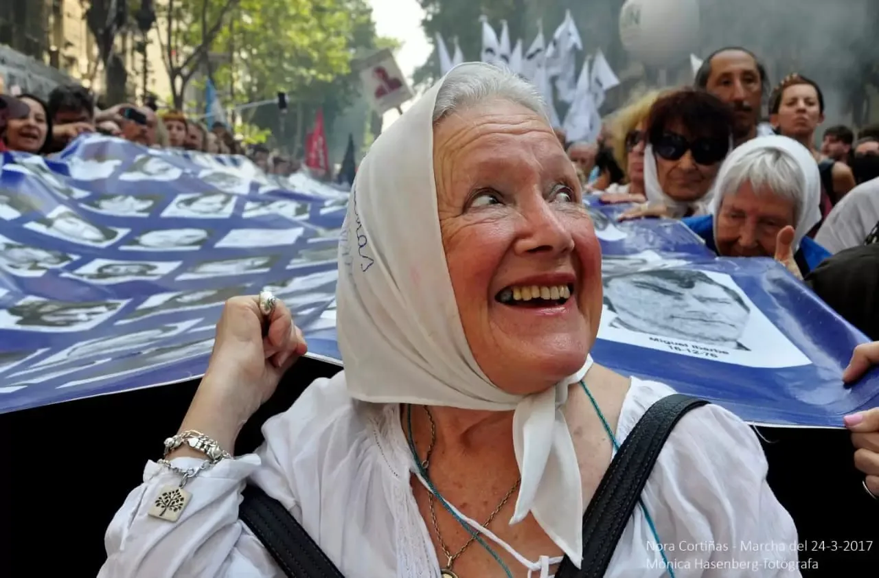 Nora Cortiñas sorri com um lenço branco na cabeça em frente a uma grande bandeira.
