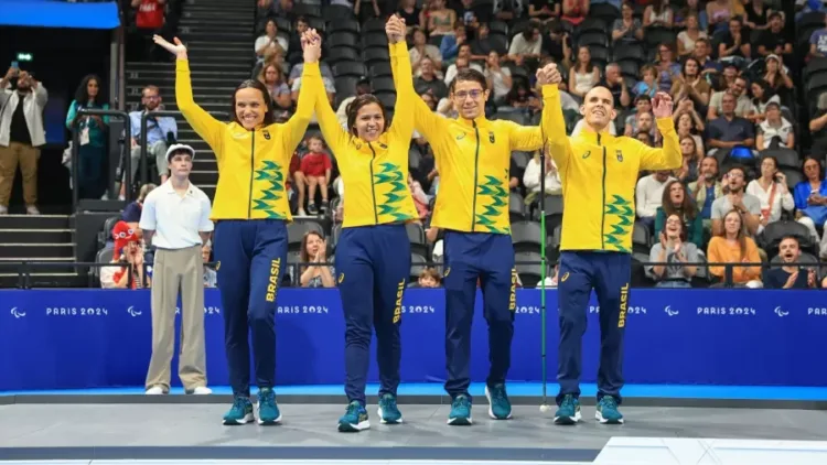 Na imagem, quatro paranadadores brasileiros celebram juntos no pódio das Paralimpíadas de Paris 2024. Da esquerda para a direita estão Carol Santiago, Lucilene Souza, Douglas Matera e Matheus Rheine. Todos vestem agasalhos amarelos e calças azuis com o nome "Brasil" estampado na perna, completando o uniforme oficial da delegação brasileira. Carol e Lucilene estão com os braços levantados, sorrindo amplamente. Douglas e Matheus também erguem os braços, com expressões de alegria e orgulho. Ao fundo, o público aplaude, enquanto os atletas celebram sua conquista.