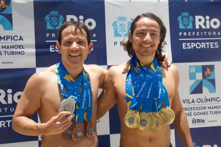 Na imagem, os irmãos Douglas e Thomaz Matera estão lado a lado, exibindo suas medalhas conquistadas no Parapan de 2023. Eles estão sem camisa, em um ambiente externo, com um fundo de banners da Vila Olímpica e da Prefeitura do Rio de Janeiro. Douglas, à esquerda, segura uma medalha de prata e usa outras medalhas no pescoço, enquanto Thomaz, à direita, exibe várias medalhas de ouro penduradas. Ambos sorriem, aparentando orgulho e alegria pelas suas conquistas.